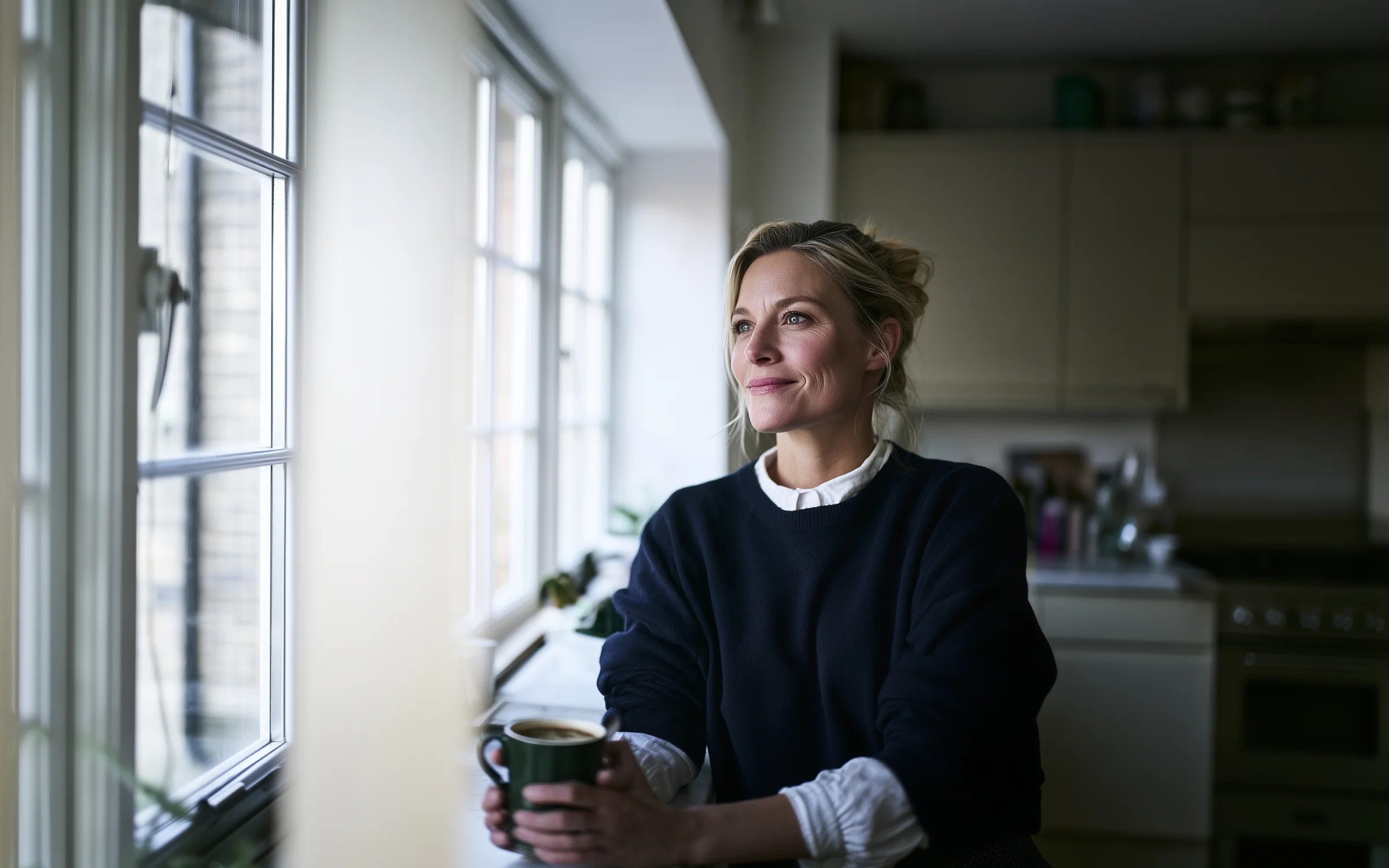 A woman with a mug of coffee looking out of her kitchen window in the early morning